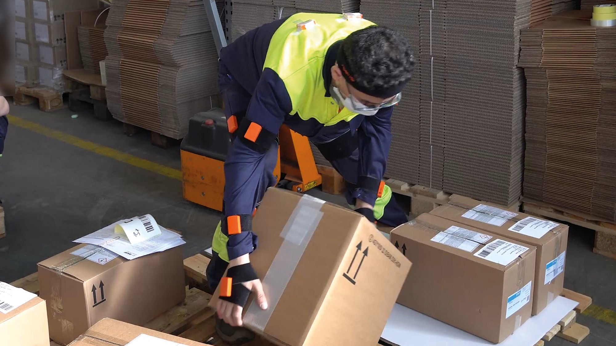 Ein Arbeiter in Schutzkleidung und mit Schutzbrille hebt einen Karton in einem Lagerbereich an. Im Hintergrund sind gestapelte Kartons und Verpackungsmaterial zu sehen. Der Arbeiter verwendet orangefarbene Sensoren an Armen und Beinen, möglicherweise zur Überwachung seiner Bewegungen oder Haltung. Eine zweite Person, deren Gesicht unscharf ist, sitzt links und überprüft Dokumente oder Geräte.