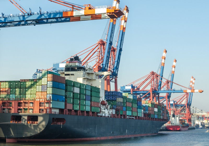 Ein großes Frachtschiff, beladen mit Containern in verschiedenen Farben, liegt in einem Hafen. Über dem Schiff ragen mehrere blaue und orangefarbene Containerkräne empor, die für das Be- und Entladen der Fracht verwendet werden. Im Hintergrund sind weitere Schiffe und Kräne zu sehen, während der klare Himmel das maritime Hafenbild abrundet.
