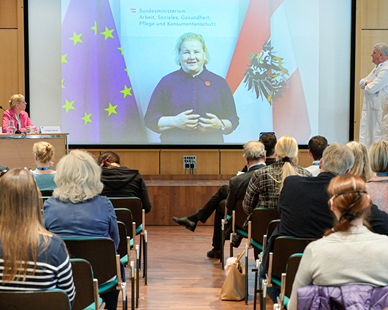 In einem Konferenzraum verfolgen mehrere Personen eine Präsentation mit einer großen Leinwand, auf der eine Person vor den Flaggen der Europäischen Union und Österreichs zu sehen ist.