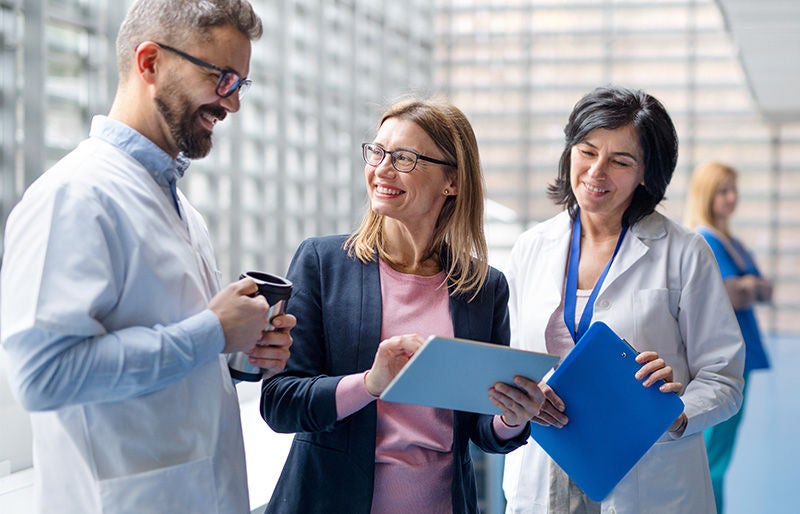 Group of doctors talking to pharmaceutical sales representative. mehrere Personen stehen beisammen, lächelnd, mit Cafetassen, Tablet bzw. einer Schreibunterlage in den Händen. Die Umgebung wirkt sehr modern; manche Personen haben einen weißen Arztkittel an
