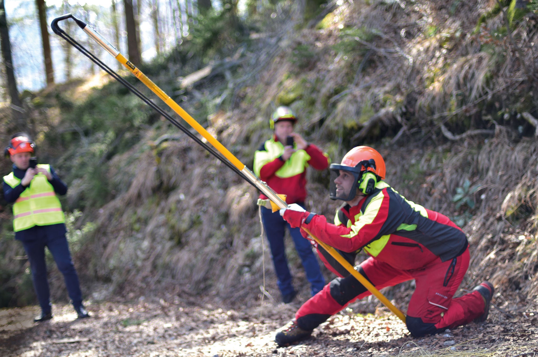eine Gruppe Arbeiter stehen im Wald, ein Arbeiter mit Helm hält eine Metall-Stange, dahinter stehen zwei Männer, die mit ihren Handys filmen