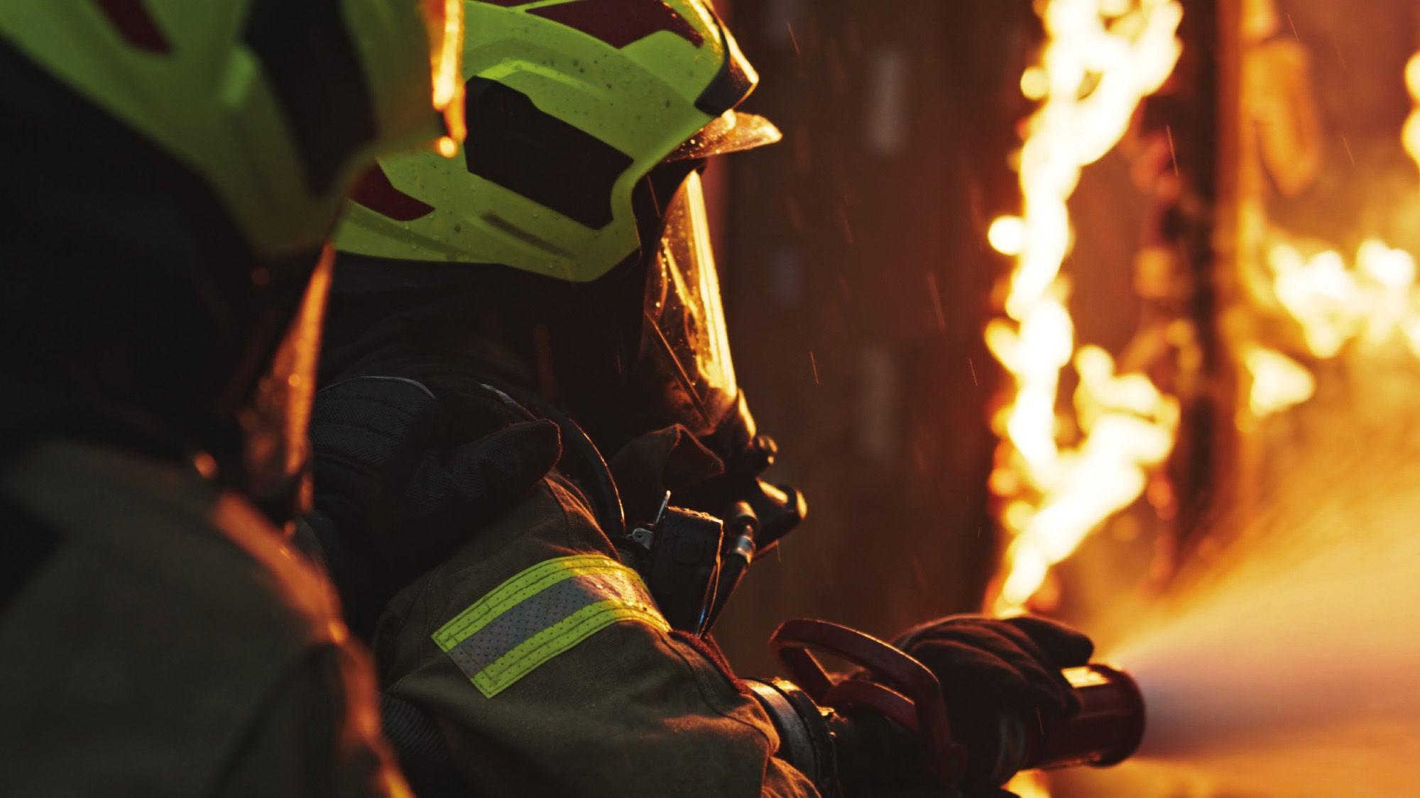 Zwei Feuerwehrleute in Schutzkleidung und Helmen kämpfen gegen ein intensives Feuer. Der eine Feuerwehrmann hält einen Schlauch, aus dem Wasser spritzt, während sie sich in unmittelbarer Nähe der Flammen befinden. Die Szenerie ist in warmen, flackernden Lichttönen gehalten, die die gefährliche Umgebung betonen.