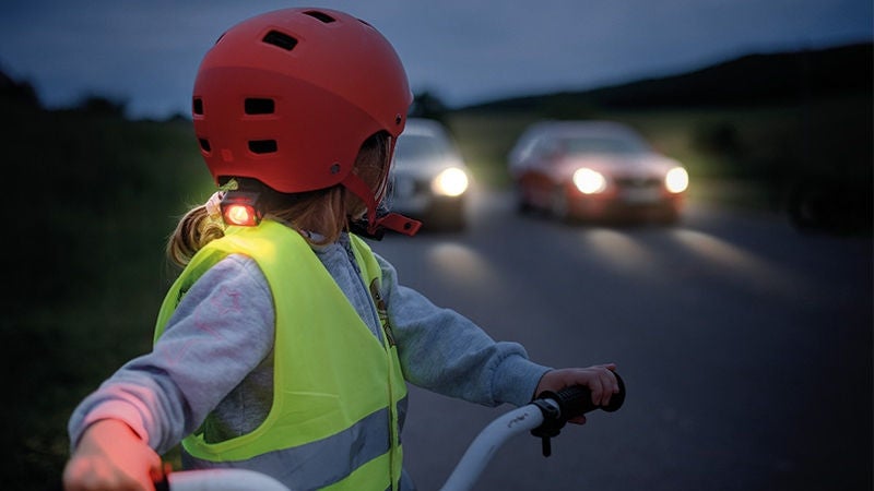 ein Mädchen fährt mit Helm, Schutzweste, Stirnlampe (im Nacken) Rad, es wirkt dämmrig, dahinter zwei Autos