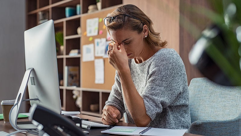Eine Person mit Brille auf dem Kopf sitzt an einem Schreibtisch vor einem Computer und hält sich erschöpft die Stirn in einem Büro mit Regalen und Pinnwand im Hintergrund.