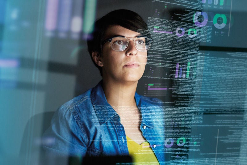 Bild einer jungen Frau mit Brille, die vor einem transparenten Bildschirm mit Datenvisualisierungen steht. Oben rechts ein Bereich mit einem unscharfen oder verpixelten Element.