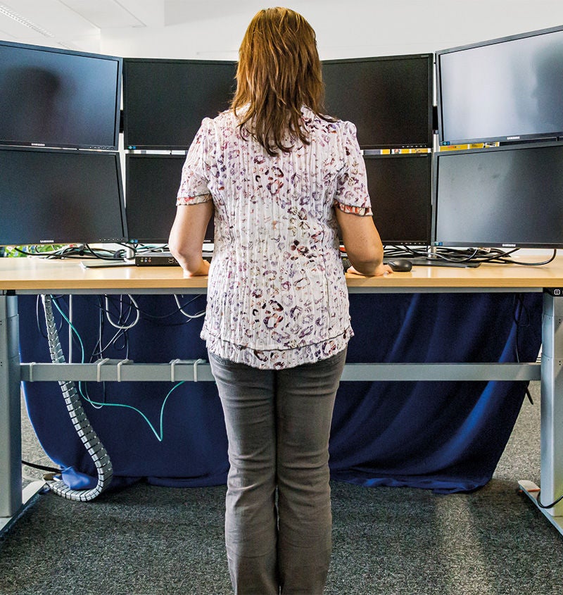 Rückenansicht auf eine Frau, die an einem Schreibtisch steht und vor sich viele Monitore hat