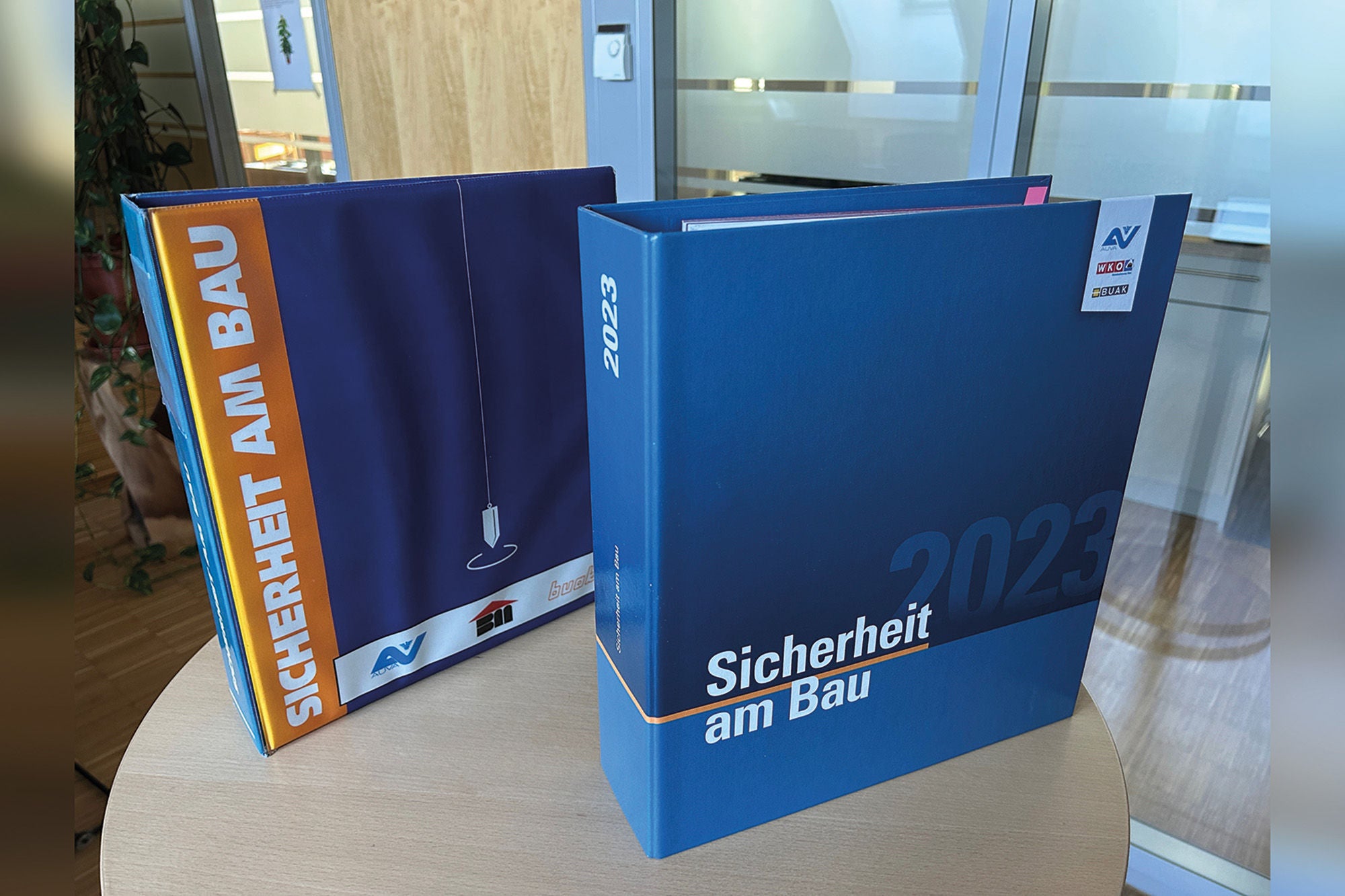Zwei breite Ordner mit der Aufschrift „Sicherheit am Bau“, einer in Blau mit Jahreszahl 2023, der andere in Lila-Orange – stehend auf einem runden Holztisch in einem Büro.