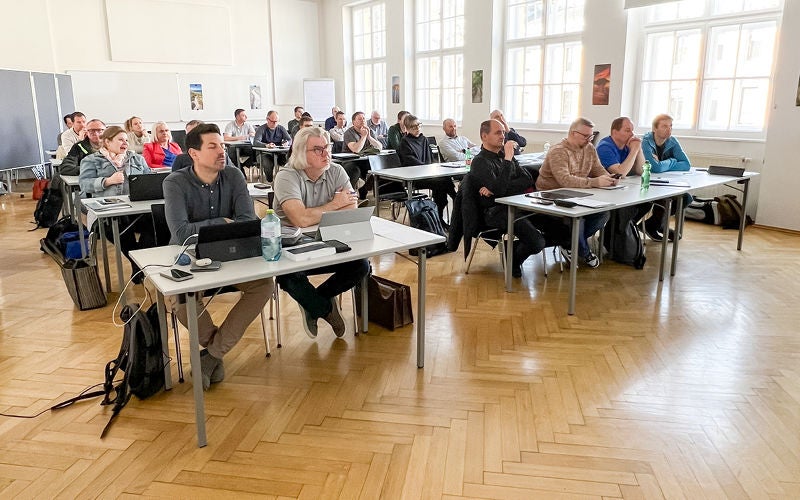 Teilnehmende sitzen an Tischreihen in einem hellen Seminarraum und verfolgen einen Vortrag.