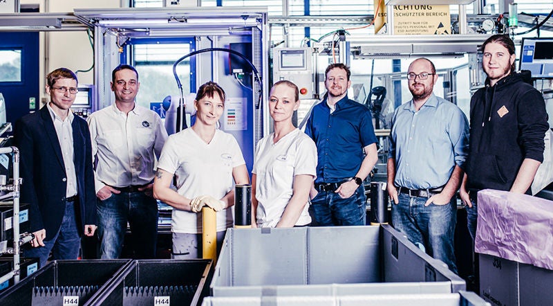 Team steht in einer Produktionsumgebung vor einer Anlage und blickt in die Kamera.