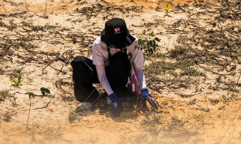 Eine Person arbeitet gebückt auf sandigem Boden und schützt sich mit langer Kleidung, Handschuhen und einer schattenspendenden Kopfbedeckung vor der Sonne.