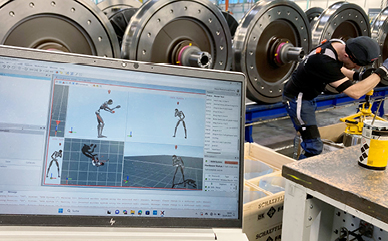 Ein Arbeitsplatz in einer Werkstatt zeigt eine Person in Arbeitskleidung, die an einer Maschine arbeitet, während im Vordergrund ein Laptop mit einer 3D-Animationssoftware geöffnet ist.