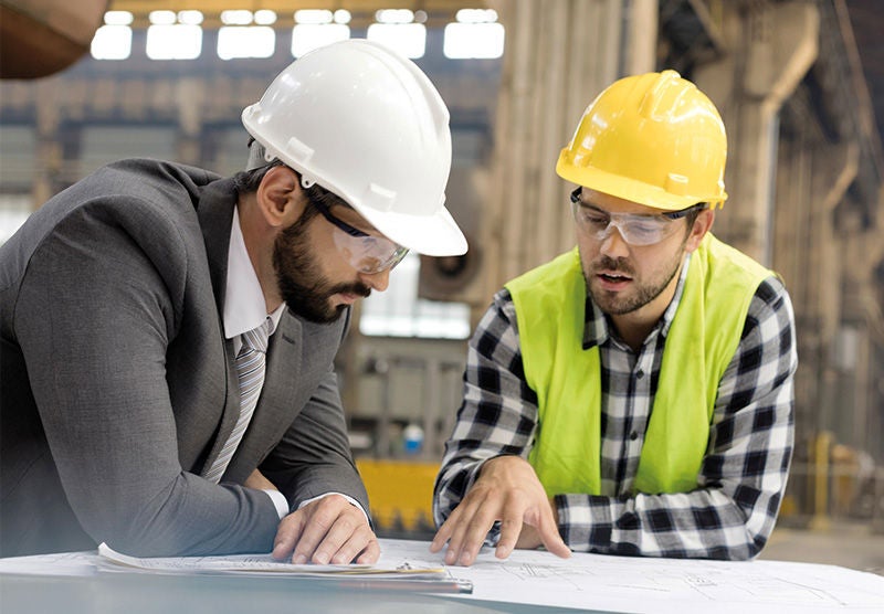 Zwei Männer mit Schutzhelm und Schutzweste stehen bei einem Schreibtisch beisammen und besprechen einen Plan