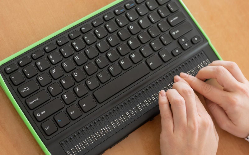 Eine Person liest mit den Fingern eine Braillezeile unterhalb einer Computertastatur, um digitale Inhalte barrierefrei nutzen zu können.