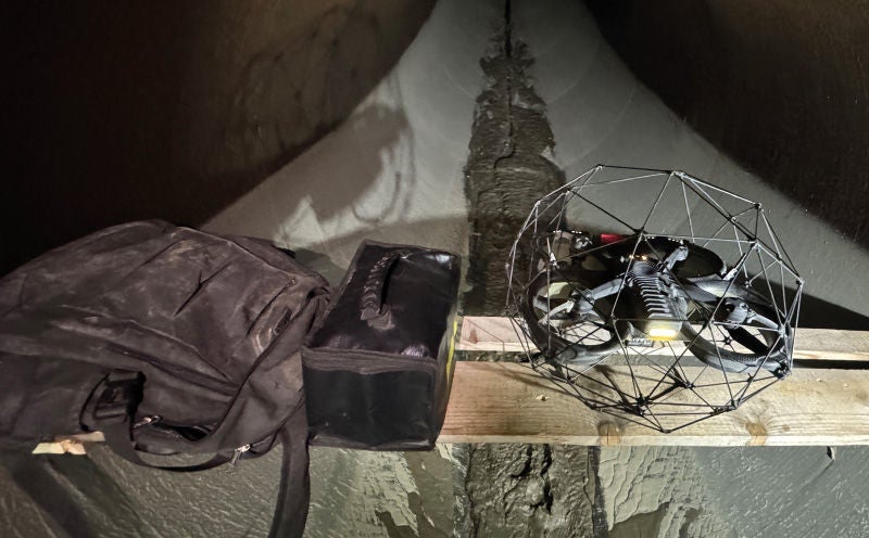Eine Drohne in einem Schutzkäfig liegt auf einem Holzbrett in einem engen Tunnel. Neben der Drohne befinden sich eine Tasche und ein Rucksack. Die Drohne ist für den Einsatz in gefährlichen oder schwer zugänglichen Bereichen der Bau- und Bergbauindustrie konzipiert.