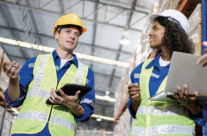 Arbeiter:innen mit Helm und Schutzweste stehen in einer Lagerhalle beisammen