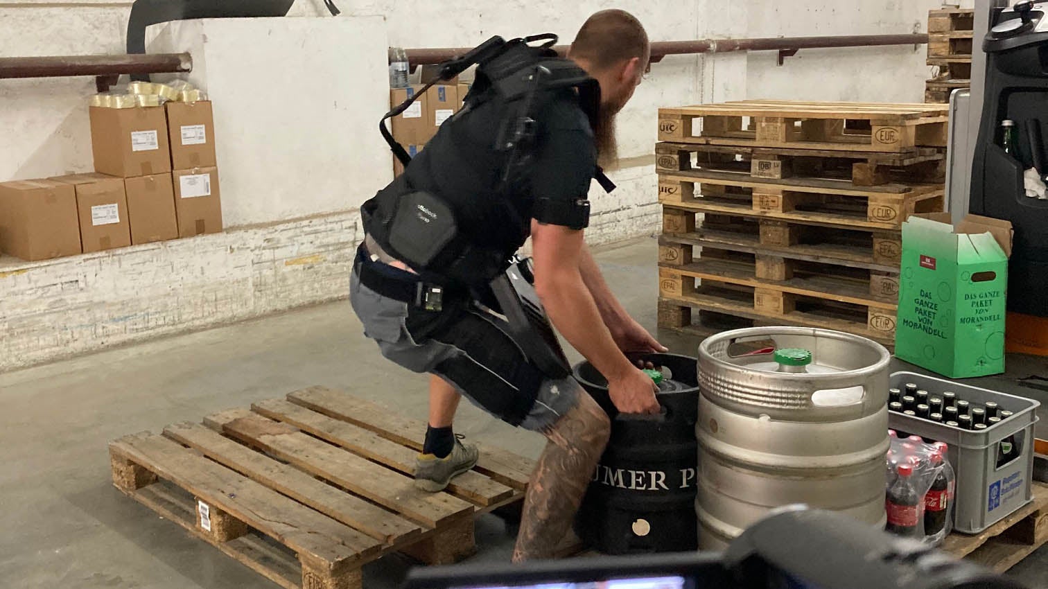 Das Bild zeigt einen Arbeiter mit angelegtem Exoskelett beim Heben eines Bierfasses vom Boden auf eine Palette. Bei dieser Tätigkeit wird er von einem Captiv-Motion-System gefilmt.