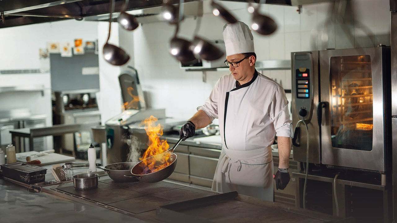 Bild zeigt einen Koch, der in der Küche eines Gastronomiebetriebes steht und eine rauchende Pfanne in der Hand hält